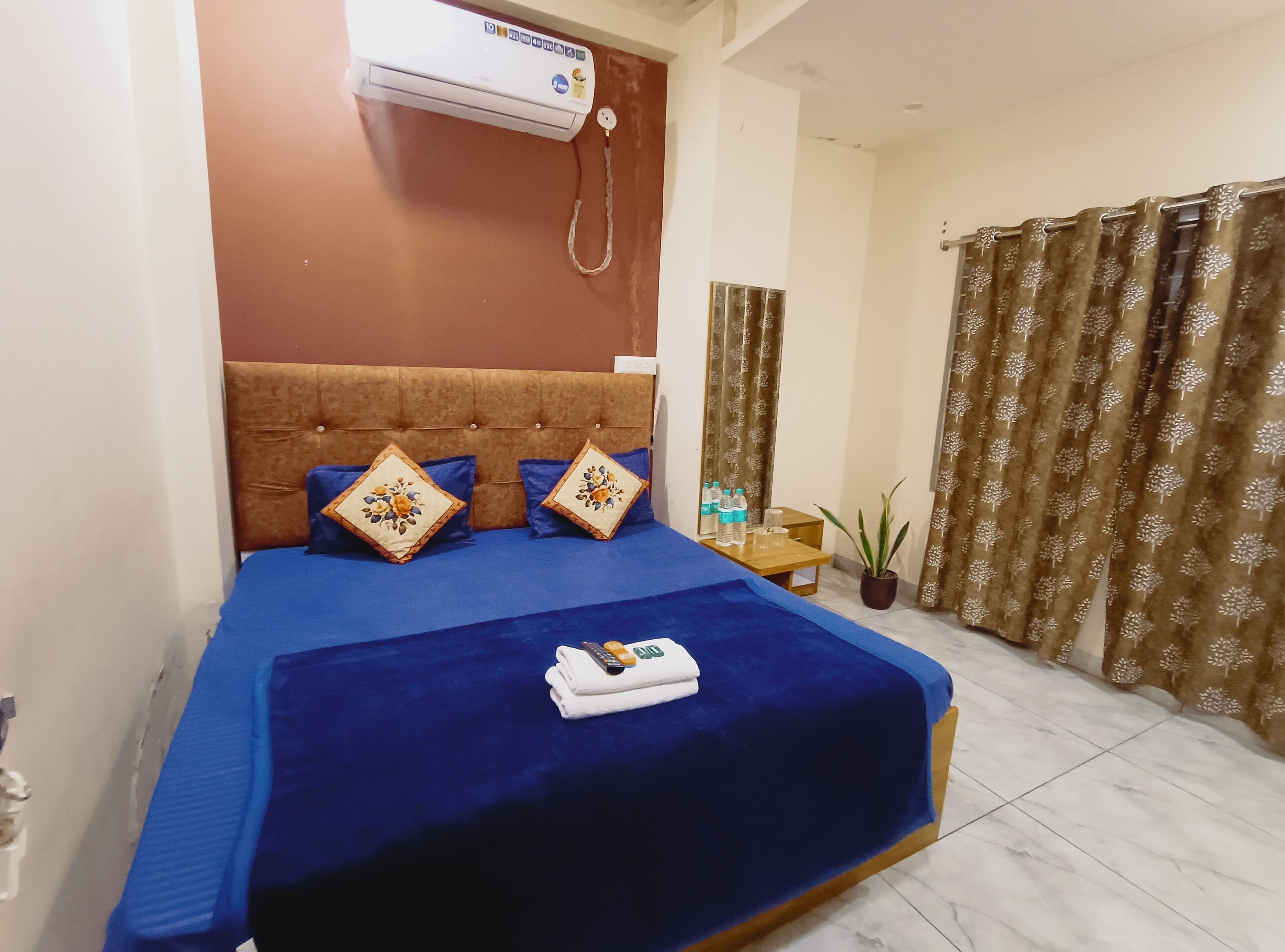 Habitación de hotel con cama, mesita de noche con botellas de agua, planta en maceta y equipo de aire acondicionado.