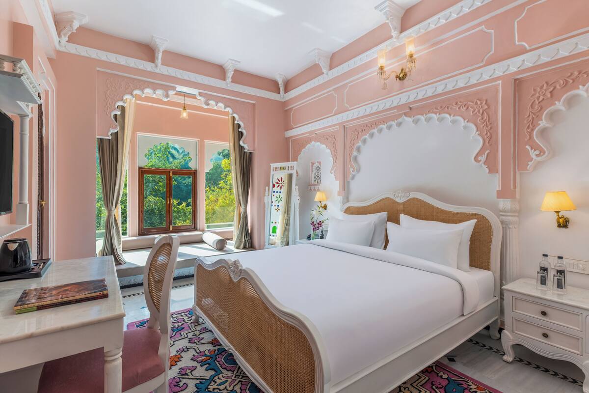 Ein Schlafzimmer mit einem großen Bett, einem Schreibtisch und einem Stuhl. Ein Fenster mit Vorhängen, ein Spiegel und eine Lampe auf dem Nachttisch sind ebenfalls vorhanden.