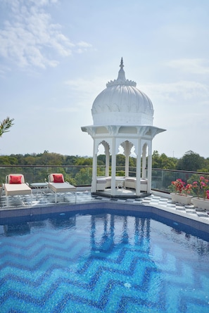 Uma piscina com padrão quadriculado, um gazebo branco e duas cadeiras de lounge com almofadas vermelhas.