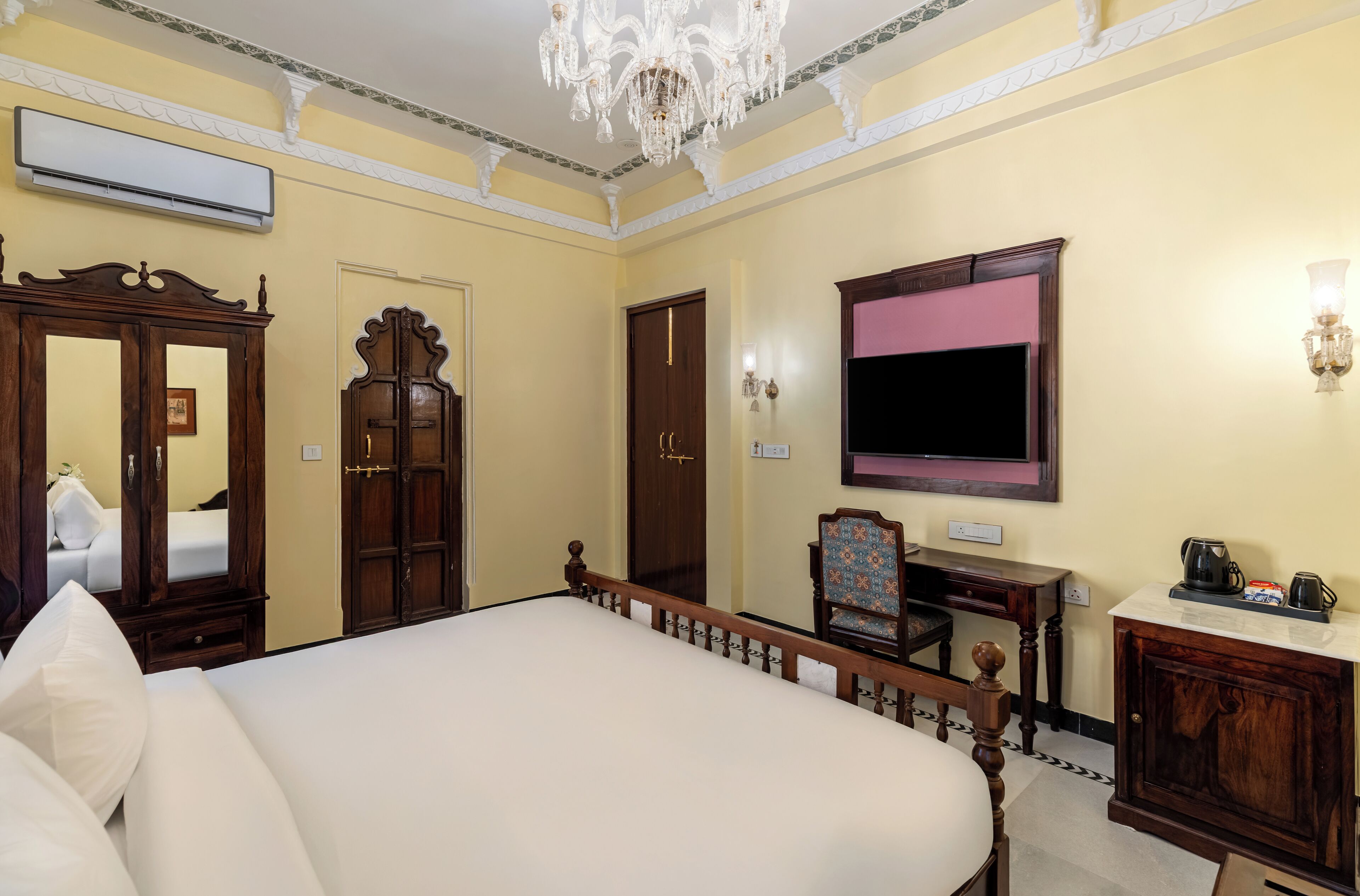A hotel room with a large bed, wooden furniture, a chandelier, a flat-screen TV, and a minibar.