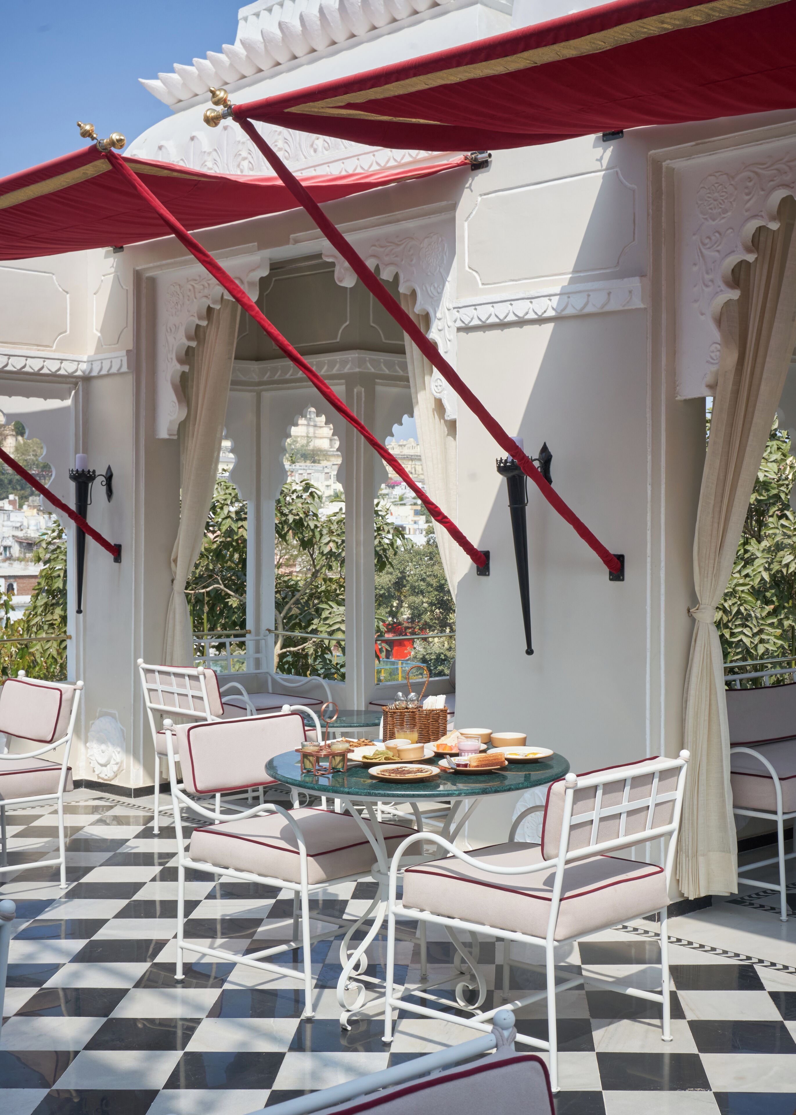 A terrace with white metal chairs and a table set for dining, featuring a checkered floor and red canopies.