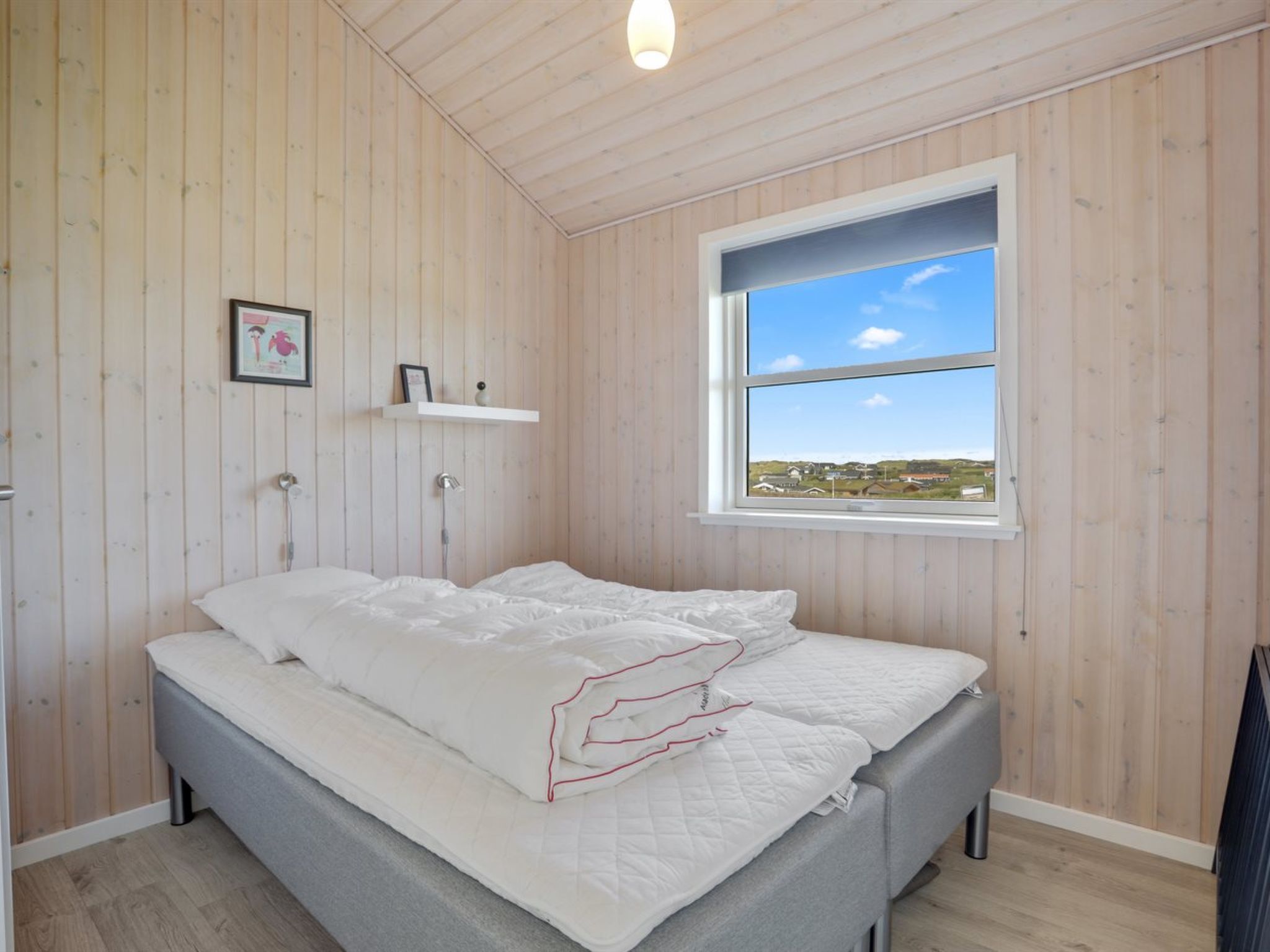 Un dormitorio con paredes de madera, una cama con ropa blanca, una ventana con vistas y una estantería con un cuadro y un florero.