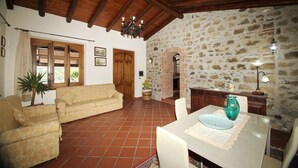 A living room with a sofa, a dining table with chairs, a chandelier, and a stone wall. - VVilla Cosentino is an enchanting retreat immersed in the Mediterranean nature of Sicily, where peace, privacy and authenticity come together. Surrounded by centuries-old olive trees, fruit trees and breathtaking sea views, this villa is the ideal place  (Acquedolci)