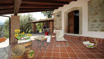 Eine überdachte Terrasse mit Backsteinwand, einem Holztisch, einem Korbwannenstuhl und einem Stuhl.