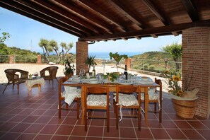 Un área de comedor al aire libre cubierta, con una mesa dispuesta para cuatro personas, rodeada de sillas de mimbre, una planta en maceta y vistas al mar.