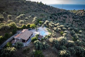 An aerial view of a property with a swimming pool, a house with a tiled roof, and a hilly landscape with sparse vegetation. - VVilla Cosentino is an enchanting retreat immersed in the Mediterranean nature of Sicily, where peace, privacy and authenticity come together. Surrounded by centuries-old olive trees, fruit trees and breathtaking sea views, this villa is the ideal place  (Acquedolci)