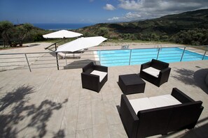 Un espace aménagé au bord de la piscine, avec des meubles d’extérieur, une piscine et une vue sur la mer et les collines.