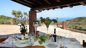 An outdoor dining table set with plates, glasses, and a bottle, overlooking a scenic landscape with hills and the sea. - VVilla Cosentino is an enchanting retreat immersed in the Mediterranean nature of Sicily, where peace, privacy and authenticity come together. Surrounded by centuries-old olive trees, fruit trees and breathtaking sea views, this villa is the ideal place  (Acquedolci)