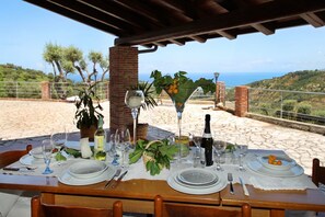 An outdoor dining table set with plates, glasses, and a bottle, overlooking a scenic landscape with hills and the sea. - VVilla Cosentino is an enchanting retreat immersed in the Mediterranean nature of Sicily, where peace, privacy and authenticity come together. Surrounded by centuries-old olive trees, fruit trees and breathtaking sea views, this villa is the ideal place  (Acquedolci)