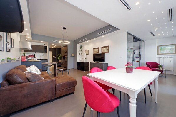 A modern living room with a brown leather sofa, a white dining table with pink chairs, and a kitchen area in the background. - Exclusive apartment on the Circus Maximus (Roma)