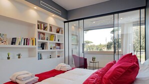 Ein modernes Schlafzimmer mit einem großen Bett, rot-weißer Bettwäsche, einem Bücherregal und Blick ins Grüne durch große Fenster.