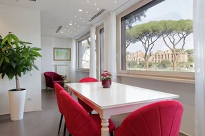 A modern dining area with a white table and red chairs, a potted plant, and a view of ancient ruins through large windows. - Exclusive apartment on the Circus Maximus (Roma)