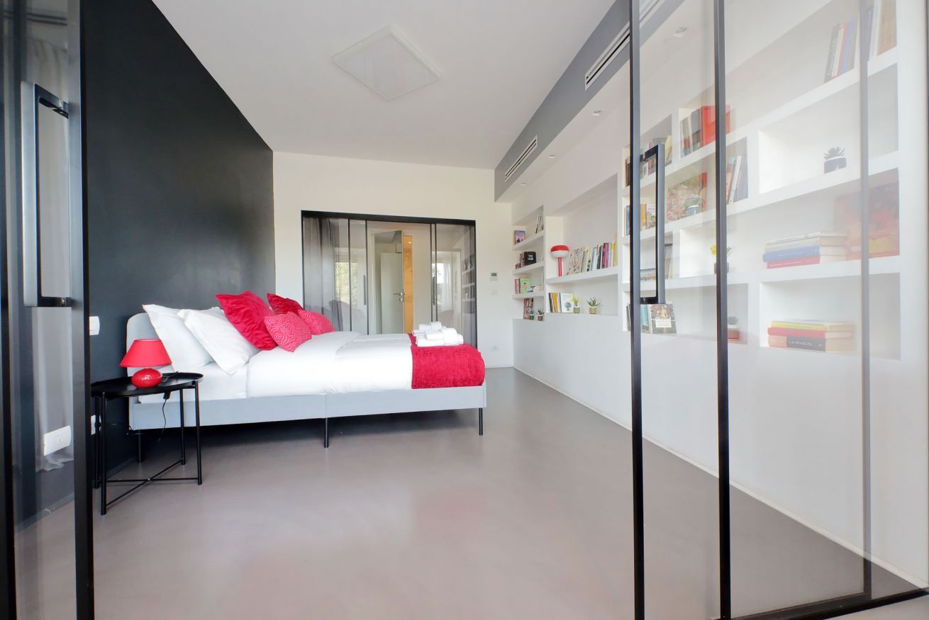 Um quarto moderno com cama branca, almofadas vermelhas e uma mesa de cabeceira com abajur.
