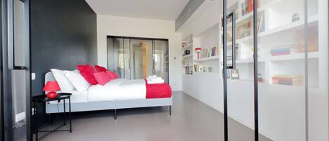 Um quarto moderno com cama branca, almofadas vermelhas e uma mesa de cabeceira com abajur.