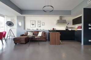 A modern living room with a brown sofa, a small coffee table, and a red chair. There is a kitchen area with dark cabinets and a hanging light fixture. - Exclusive apartment on the Circus Maximus (Roma)