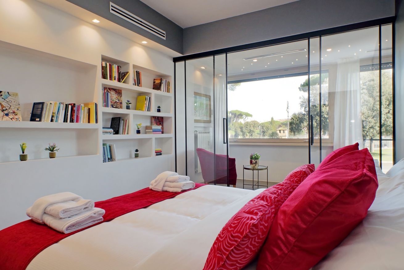 A modern bedroom with a large bed, red and white bedding, a bookshelf, and a view of greenery through large windows.
