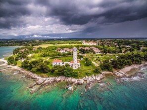 An aerial view of a coastal town with a prominent lighthouse, clear turquoise waters, and a mix of residential and natural landscapes. - Peaceful Flat Surrounded by Nature (Umag)