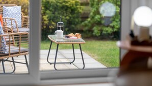 Et glastop med kaffekande, kop og bagværk på en terrasse med fletstole og udsigt til en have.