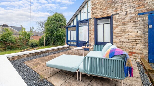 En terrasse med en lyseblå sofa og et lille bord, en murbygning med store vinduer og en blå dør.
