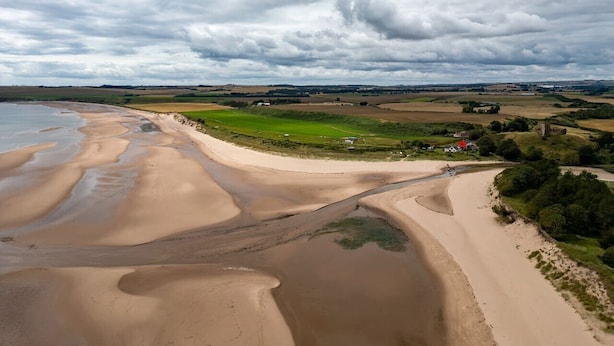 Un paysage côtier avec une plage de sable, des champs couverts d’herbe et quelques bâtiments visibles au loin.