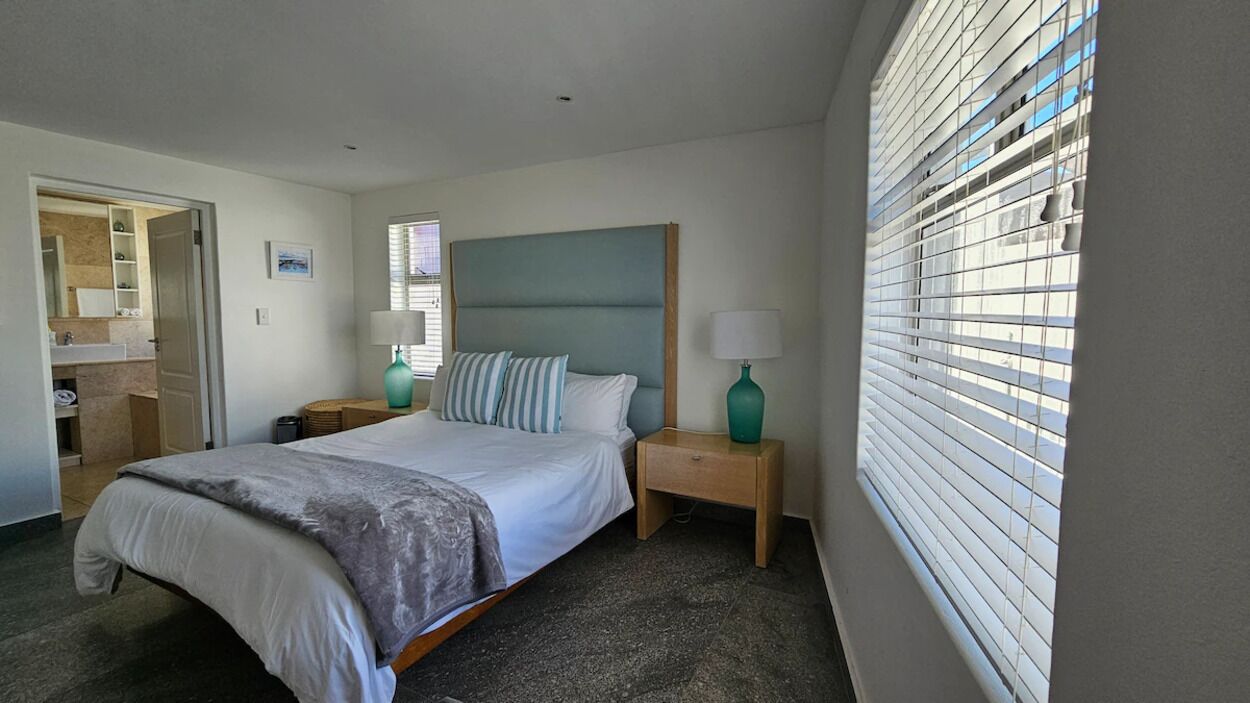 Une chambre à coucher comprenant un lit, des tables de chevet, une commode, une fenêtre avec des stores, et une salle de bain visible à travers une porte ouverte.