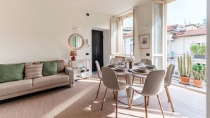 A modern living room with a sofa, dining table, chairs, and a large window with a view of buildings and cacti. - Casa Sara - MyHomeinComo (Como)