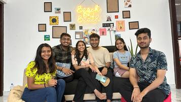 Un grupo de personas sentadas en un sofá en una habitación con una pared decorada con varias fotos enmarcadas y un letrero de neón que dice 'Beach or Beach'.