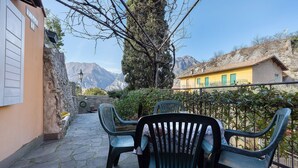 A stone-paved outdoor area with plastic chairs and a yellow building with green shutters. - Villa G with Lake View, Private Terrace Steps from Lake Garda (Torbole)