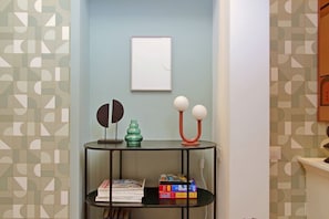 A corner of a room with a geometric-patterned wall, a framed picture, a black shelf with books and decorative items, and a small table with a green vase and a spherical object. - Lussuoso appartamento vicino al Colosseo (Roma)
