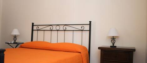 A single bed with an orange bedspread, a black metal headboard, two bedside tables with lamps, and a wooden nightstand.