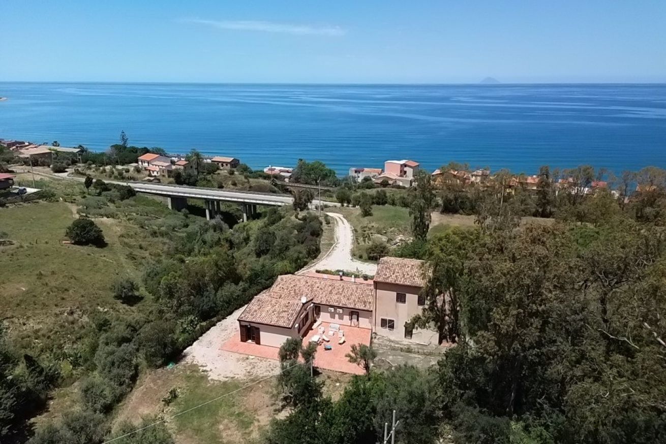 A coastal property with a paved driveway leading to a building with a tiled roof, surrounded by greenery and overlooking the sea.