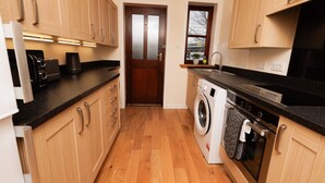 A compact kitchen with wooden cabinets, a washing machine, and a countertop with appliances. - NEW to VRBO: Central Lerwick 2Bed Apt · Garden|Walk to Sights (Lerwick)