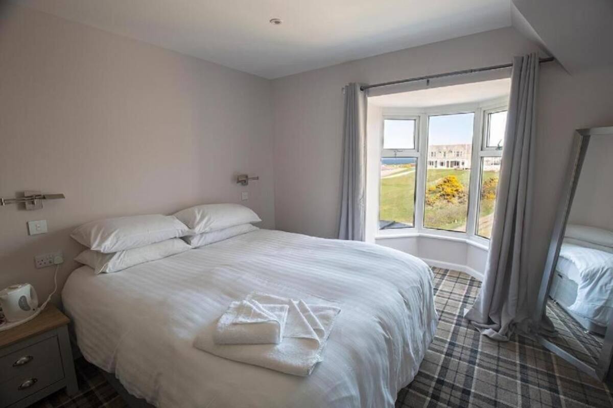 Quarto de hotel com uma cama grande, uma mesa de cabeceira com luminária, uma janela com cortinas e vista para o exterior.