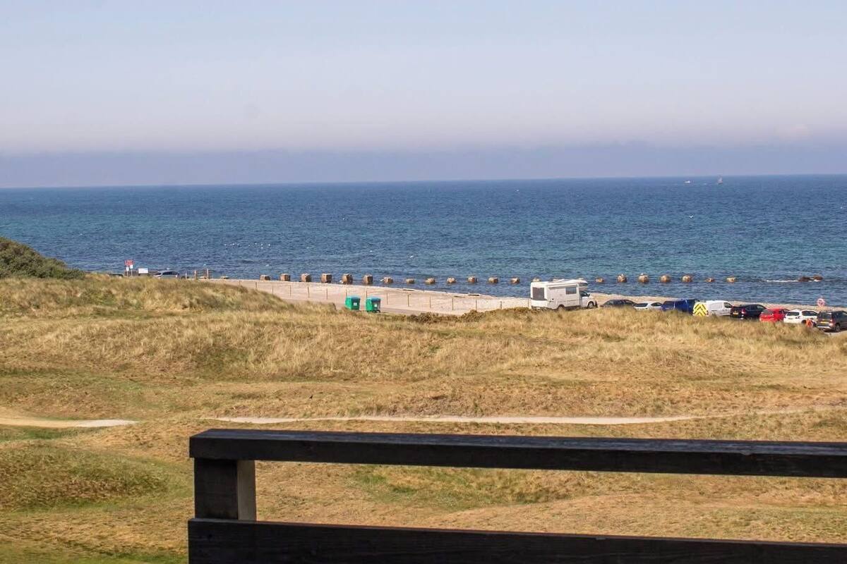 Vista costeira com uma praia de areia, veículos estacionados e um banco de madeira em primeiro plano.