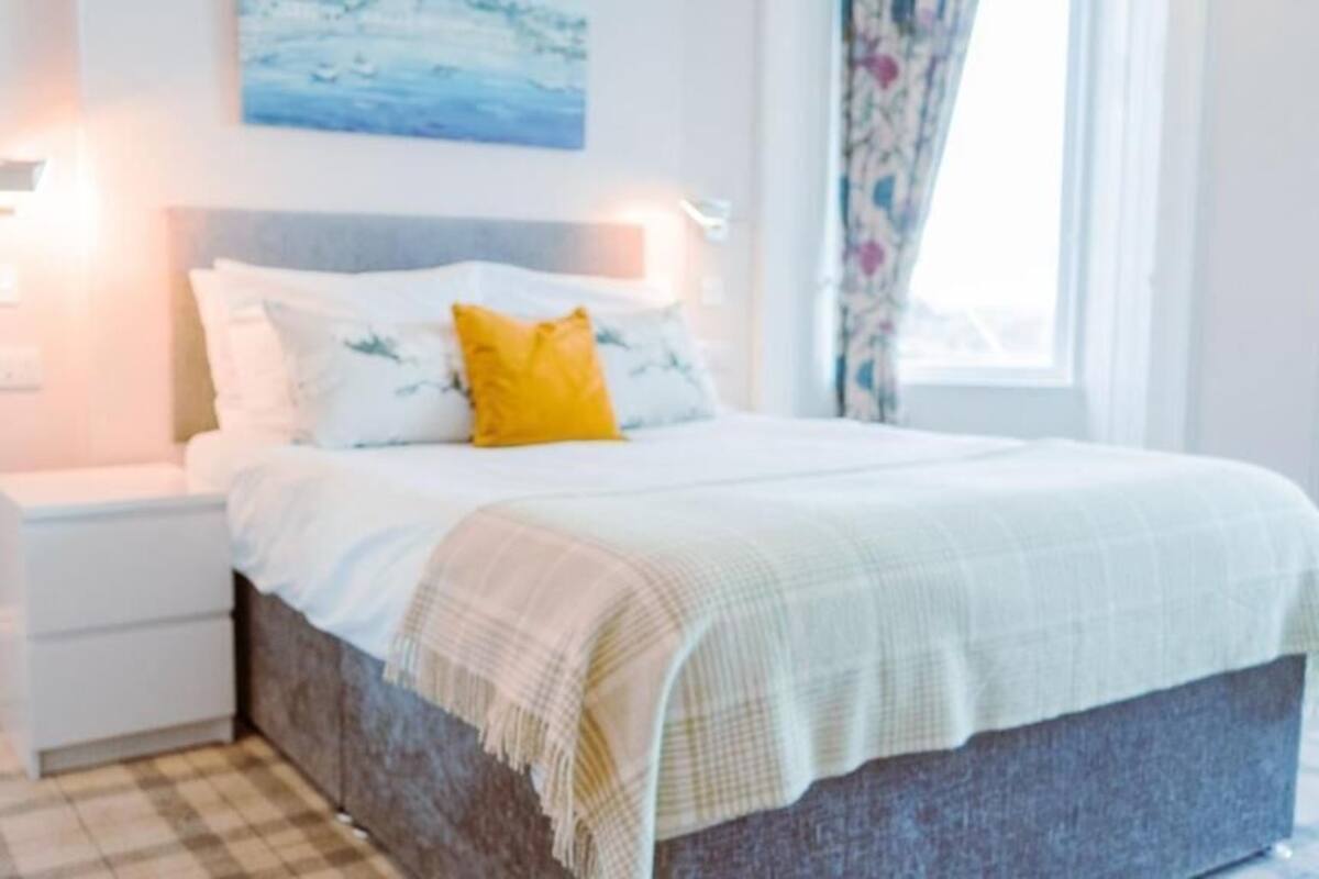 Quarto com cama, mesas de cabeceira, luminária na parede, janela com cortinas e um quadro sobre a cama.