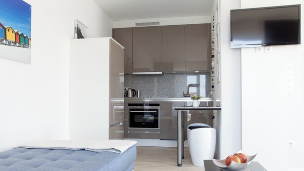 A compact kitchen with modern appliances, a dining area with a small table and chairs, and a bed in the foreground. - Refugium 1 (Emsdetten)