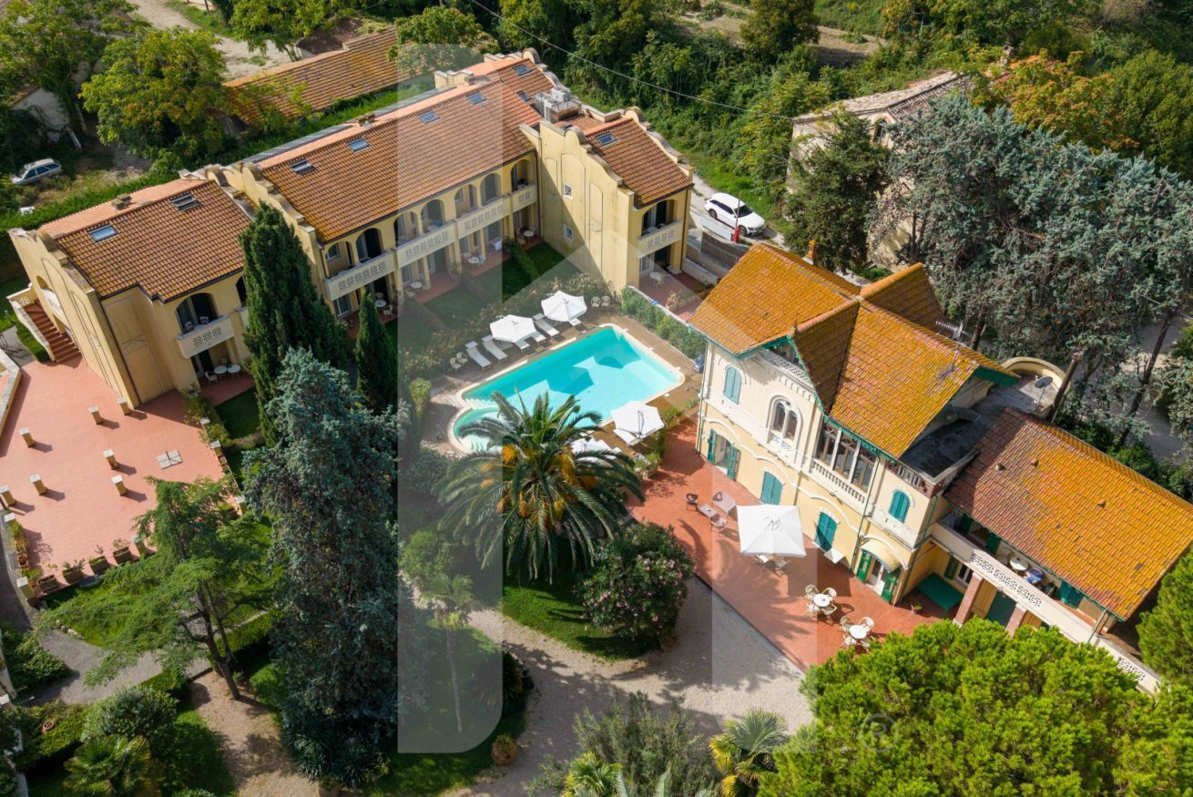 An aerial view of a hotel with a central swimming pool, multiple buildings, and surrounding greenery.
