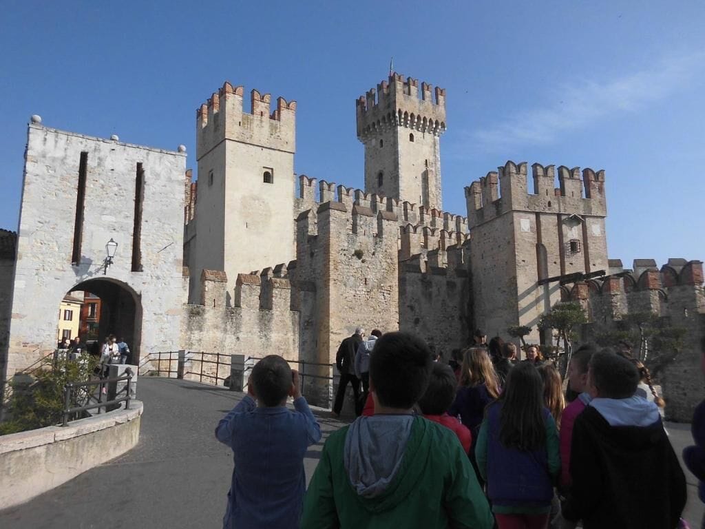 A historic stone castle with multiple towers and a central gate, surrounded by a paved walkway.