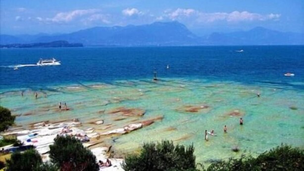 A coastal scene with clear turquoise waters, a boat in the distance, and people swimming and sunbathing on the beach. - B&B Topaze (Sirmione)
