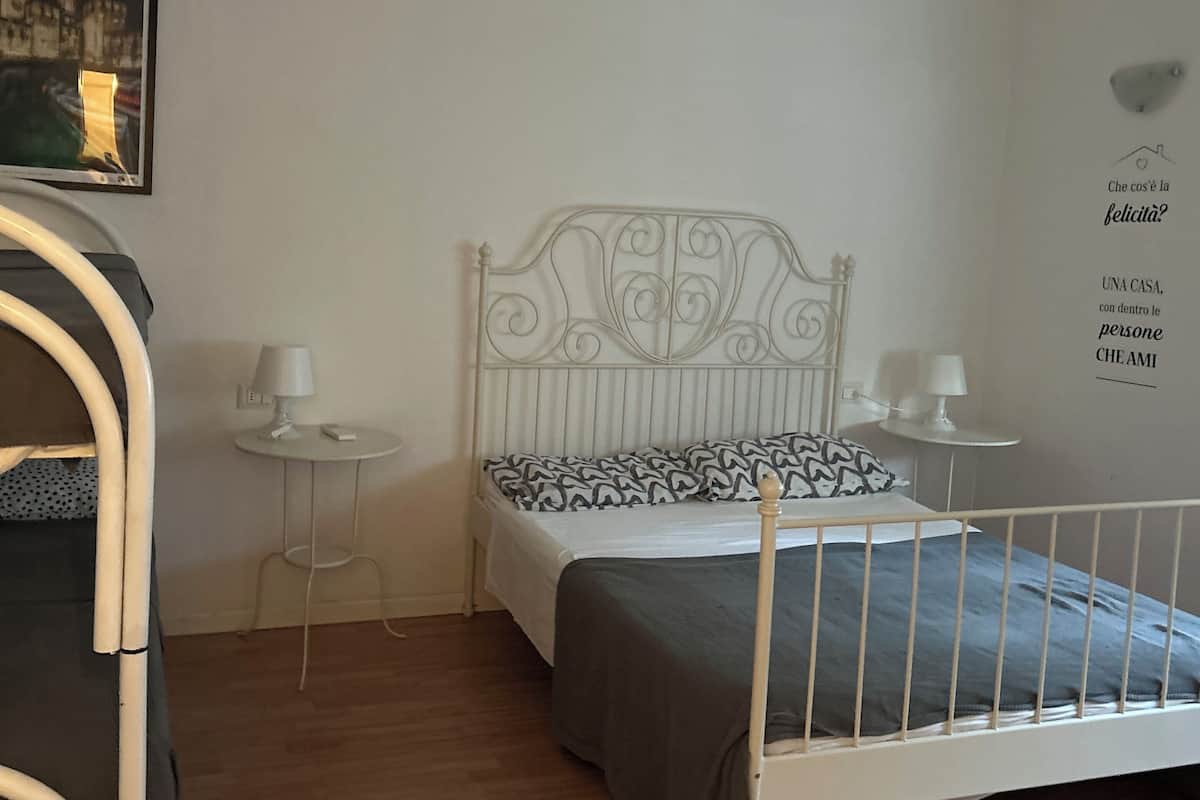 Een slaapkamer met een metalen bed, een nachtkastje met een lamp, een klein rond tafeltje met een lamp, en een muur met decoratie en tekst.