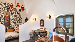 A cozy room with a stone wall, a bed with orange pillows, a chandelier, a wooden table with a copper faucet, and a view through a window. - Historical Villa Santa Croce (Santorini)