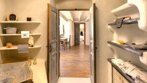 A narrow hallway with wooden floors, open doors leading to a dining area, and shelves with towels and toiletries. - Historical Villa Santa Croce (Santorini)