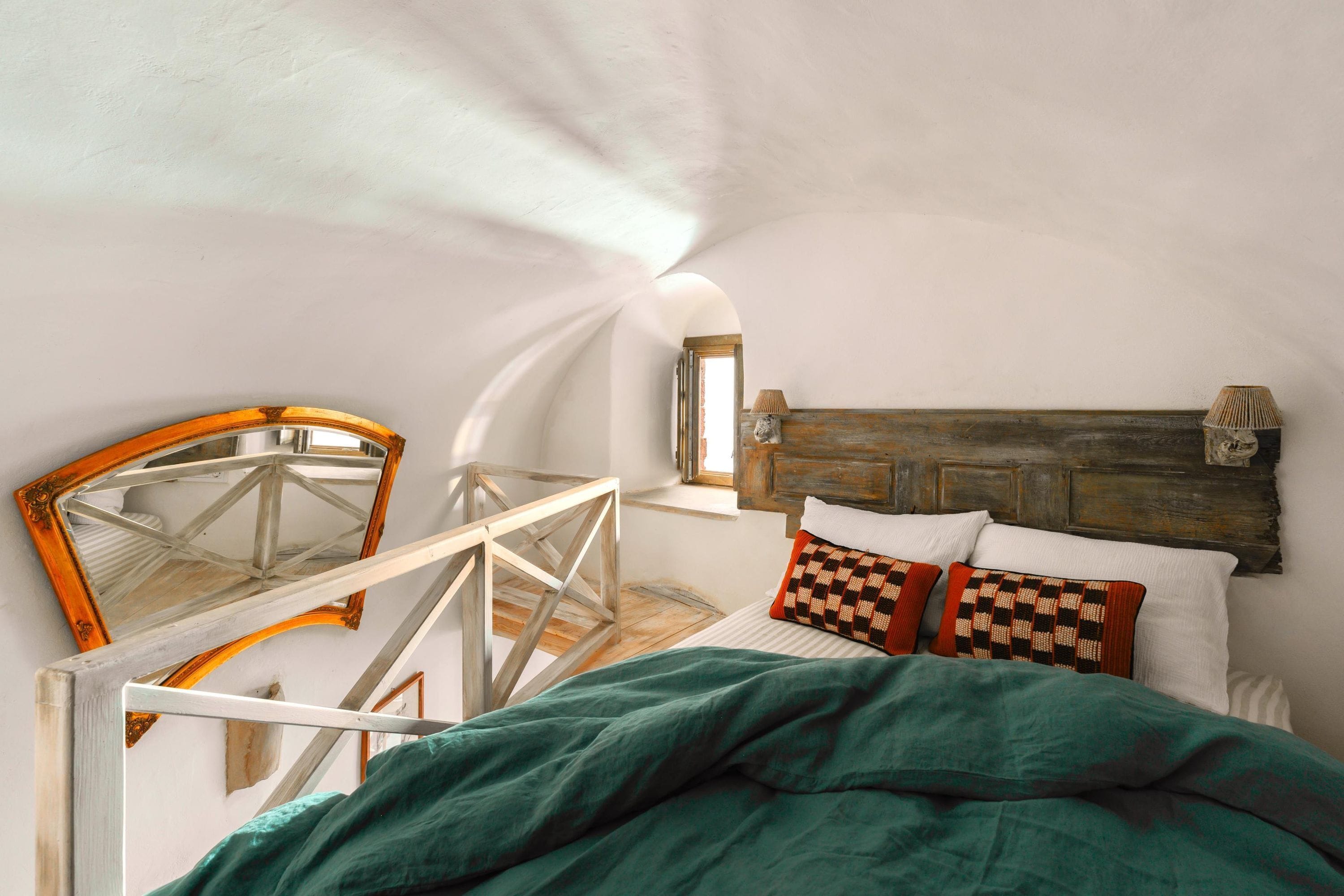 A bedroom with a bed, pillows, a wooden headboard, a window, and a mirror.