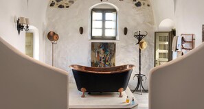 A vintage bathtub in a rustic bathroom with a large window, a painting, and a rustic lamp. - Historical Villa Santa Croce (Santorini)