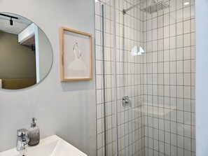 Egy fürdőszoba mosdókagylóval, tükörrel, képkeretbe foglalt műalkotással és csempézett falú zuhanyzóval.