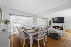 A spacious living room with a fireplace, a dining area with a wooden table and white chairs, and a large flat-screen TV mounted on the wall.