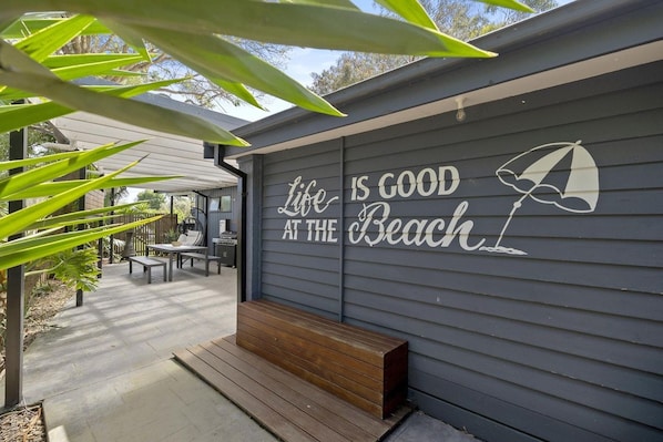 A covered outdoor seating area with a bench and a table, a wall with a motivational quote, and a view of a residential area in the background.