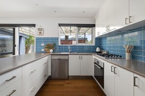 A modern kitchen with white cabinets, a stainless steel dishwasher, and a blue-tiled backsplash. - Ohana Hale, Safety Beach. (Safety Beach)