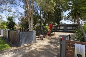 A gated entrance with a wooden fence, a red sculpture, and a building with a modern design in the background. - Ohana Hale, Safety Beach. (Safety Beach)
