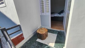 Un petit balcon étroit, avec un tabouret en bois, une rambarde en métal et une porte entrouverte donnant sur une chambre à coucher équipée d’un lit et d’une fenêtre avec des stores.